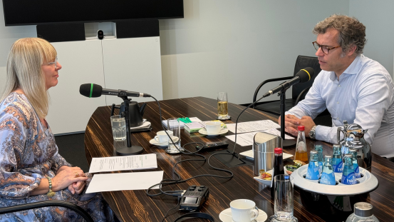Nikola Steinbock im Interview mit Dr. Andreas Möller. Beide sitzen an einem Tisch, auf dem Mikrofone, Aufnahmegeräte und Getränke stehen. Im Hintergrund steht ein Regal, ein Fernseher hängt an der Wand.