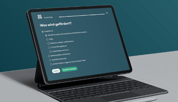 Ein Tablet mit Tastaturhülle steht auf einem Tisch. Auf dem Bildschirm ist eine Webseite mit dem Titel 'Was wird gefördert?' zu sehen. Darunter befinden sich mehrere Auswahlkästchen mit verschiedenen Fördermöglichkeiten, wie Kraftfahrzeuge, Ställe, private Wohngebäude und weitere. Am unteren Bildschirmrand sind zwei grüne Schaltflächen mit den Beschriftungen 'Zurück' und 'Ergebnisse anzeigen' sichtbar. Der Hintergrund ist einfarbig in dunklem Türkis gehalten.